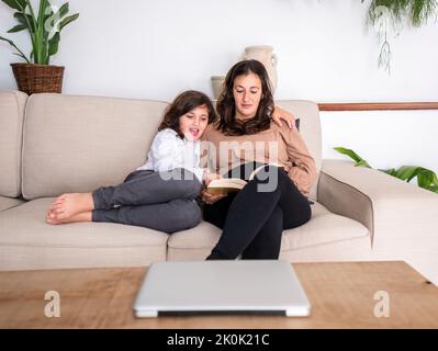 Mother reading interesting book with daughter while sitting on soft sofa in apartment with potted green plants Stock Photo