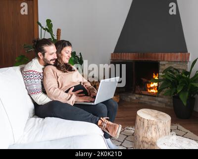 Side view full body of loving couple using laptop while sitting together on sofa placed near fireplace in modern apartment Stock Photo