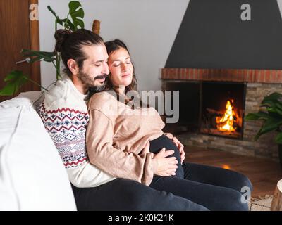 Male and pregnant female cuddling and sitting with closed eyes on comfortable sofa near fireplace in living room Stock Photo