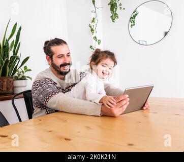 Bearded male in sweater sitting at table with daughter using tablet in light apartment with potted plants Stock Photo
