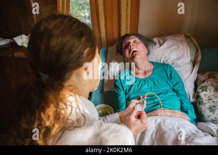 Female nurse feeding paralyzed elderly lady with puree through tube and syringe while taking care of patient at home Stock Photo