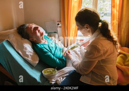 Female nurse feeding paralyzed elderly lady with puree through tube and syringe while taking care of patient at home Stock Photo