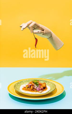 Artificial hand with red hot chili pepper levitating over plate with Mexican chicken salad on blue and yellow background Stock Photo