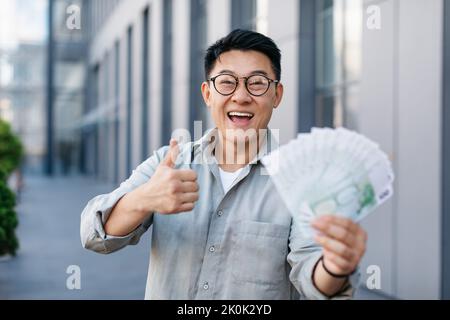 Overjoyed asian businessman holding fan of euro money and showing thumb up gesture, standing outdoors Stock Photo