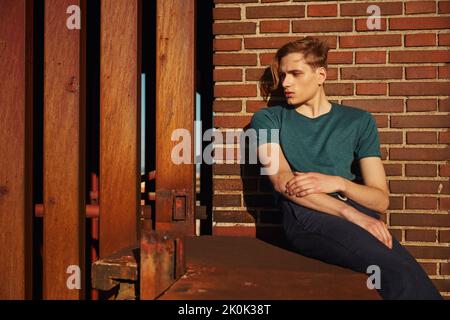 Cute young blond male resting looking away sitting on edge of roof leaning against brick wall on sunny day Stock Photo
