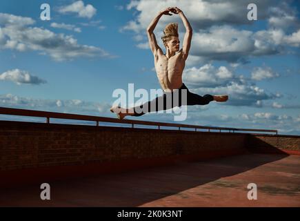 Masterful young blond ballet dancer in black leotard performing exciting jump with raising hands up on roof of building on sky background in sunny day Stock Photo