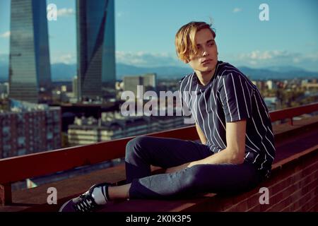 Fashionable young blond man with long bangs sitting on edge of roof of building overlooking city near fence and looking away in sunny day Stock Photo