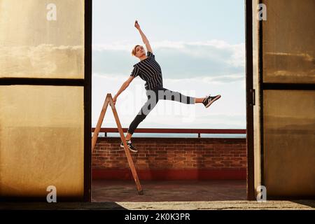 Fashionable elegant young performer standing on one leg on stepladder and raising hand to sky on roof of building on sky background Stock Photo