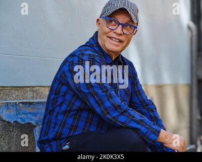 Senior male wearing trendy eyeglasses and blue plaid shirt relaxing on shabby bench on street and looking away Stock Photo