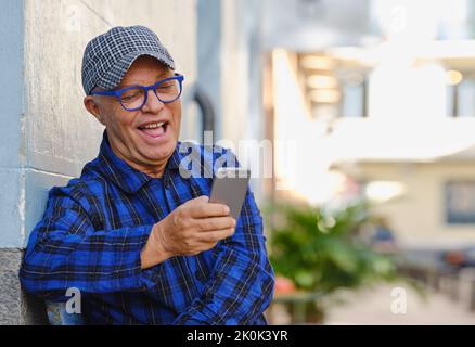 Handsome male in checkered shirt and stylish eyeglasses sitting on bench in city and browsing cellphone Stock Photo