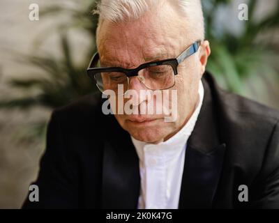Elderly male in casual wear sitting at table in cozy cafeteria and looking down Stock Photo