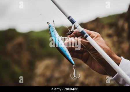 Crop anonymous male with special blue plug on fishing rod with hook standing in nature on blurred background during fishing Stock Photo