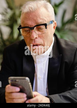 Elderly male in casual wear sitting at table in cozy cafeteria using smartphone Stock Photo