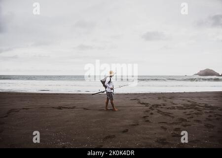 Full body side view of anonymous man with fishing rod with rucksack strolling on sandy shore near sea in nature Stock Photo