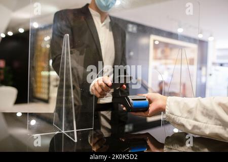 Crop anonymous male buyer in protective mask using smartphone to make contactless payment at reception desk with protective glass screen Stock Photo