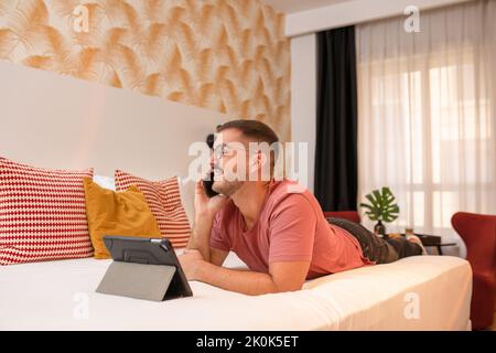 Side view of positive bearded male in eyeglasses talking on the mobile phone next to tablet while lying on bed in hotel room Stock Photo