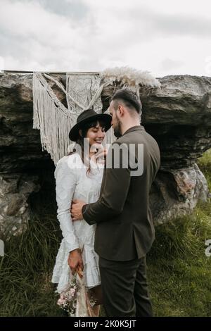 Delighted newlywed couple in stylish clothes standing against macrame hanging on rock in nature on wedding day and embracing Stock Photo