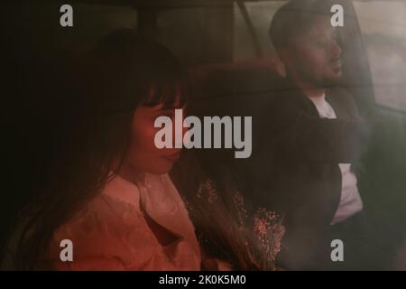 View of charming bride in white dress and with floral bouquet sitting in car with red neon light against groom on wedding day Stock Photo