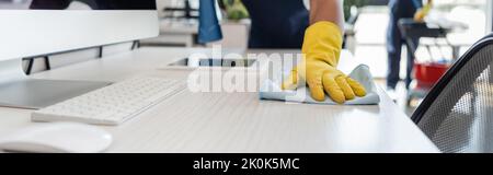 cropped view of cleaner in rubber glove washing desk near computer monitor and digital tablet, banner,stock image Stock Photo