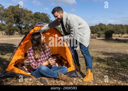 Young female browsing on mobile phone while boyfriend prepares camping tent in forest Stock Photo