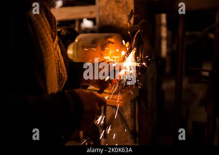 Unrecognizable elderly craftsman using bench grinder to shape metal part while working in dark workshop Stock Photo