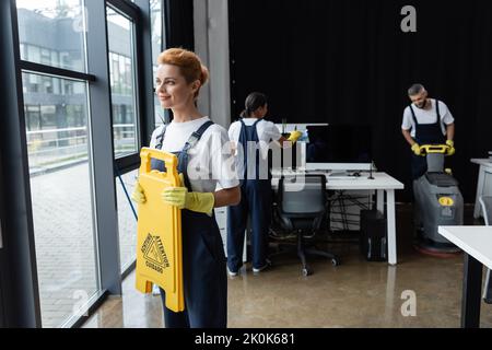 smiling woman holding attention board near windows while interracial colleagues cleaning office,stock image Stock Photo
