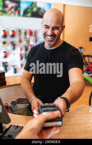 Delighted adult man in black t-shirt smiling and giving payment terminal to crop client while working in bike shop Stock Photo