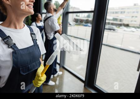 smiling woman in overalls holding window squeegee near blurred multiethnic colleagues,stock image Stock Photo
