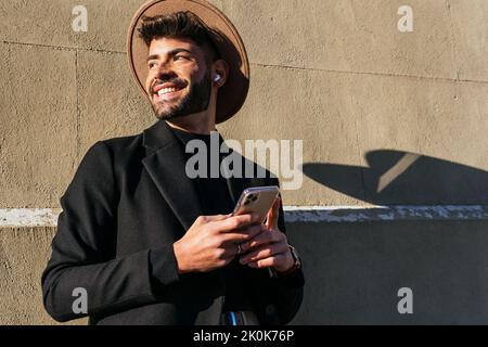 Young content unshaven male in formal apparel with cellphone and wireless earbud looking away in city in sunlight Stock Photo