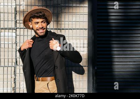 Young smiling masculine male in formal outfit with wireless earbuds looking away while strolling in town on sunny day Stock Photo