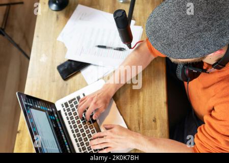 From above unrecognizable male worker with headphones listening to audio while surfing netbook and reading papers at table in modern studio with profe Stock Photo