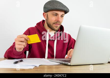 Bearded male worker with credit card browsing on laptop while paying with credit card online at desk with documents during work in office Stock Photo