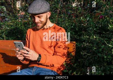 Positive male in cap surfing internet on cellphone while sitting on bench in park near green plants on sunny street Stock Photo
