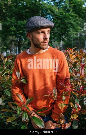 Calm bearded male in headdress looking away while standing among lush shrub in park against tall green trees in city Stock Photo