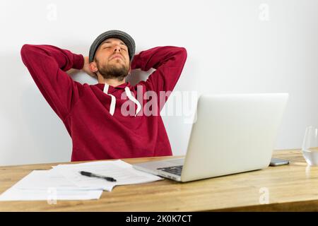 Male employee with closed eyes with hands behind head sitting at table with netbook and documents during work in office Stock Photo