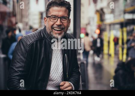 Smiling handsome middle aged man in glasses and leather jacket looking at camera while standing in urban street Stock Photo