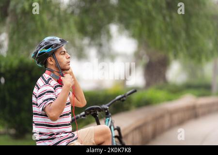 Latin male in casual wear putting on helmet while sitting on stone border near bicycle in city with trees Stock Photo