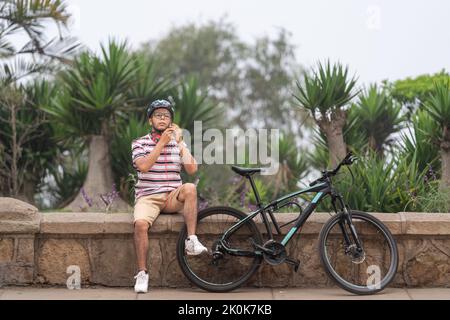 Full body of Latin male in casual wear putting on helmet while sitting on stone border near bicycle in city with trees Stock Photo