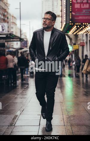 Confident thoughtful handsome middle aged man in glasses and leather jacket looking up while walking in urban street Stock Photo