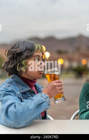 Delighted young woman sitting at table in veranda of pub in city of Cusco and drinking glass of beer and looking away Stock Photo