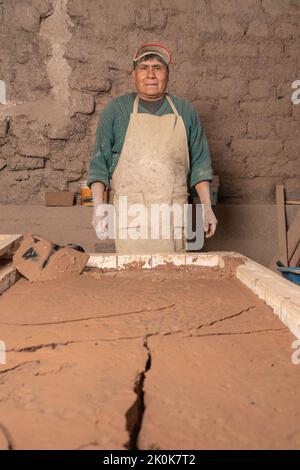 Calm Peruvian male worker with dirty hands in uniform looking at camera while standing near plaster near wall during work in ceramics workshop Stock Photo