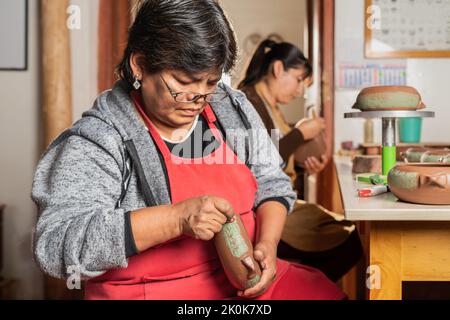 Concentrated Peruvian female artisan in apron painting ceramic bowl with green color on stand while working in light professional workshop Stock Photo