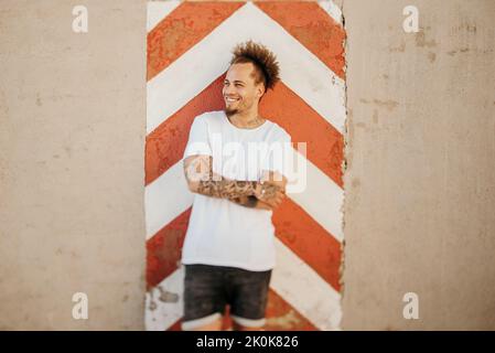 Positive young ethnic man in casual clothes smiling and looking away while leaning on weathered wall on city street Stock Photo