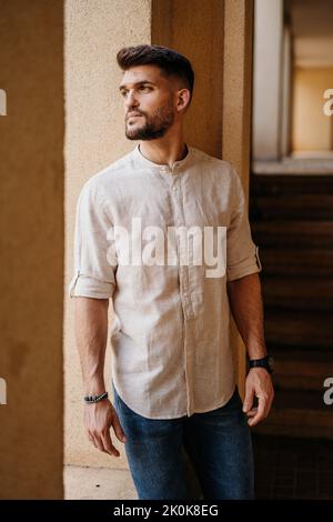 Low angle of serious confident unshaven male in light shirt and jeans looking out window with interest and thinking while standing leaning on wall aga Stock Photo