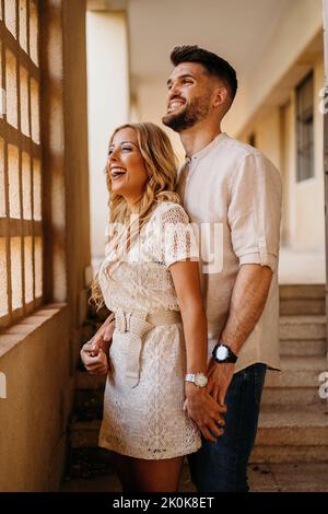Handsome bearded man and gorgeous positive woman in light dress looking out window and smiling while standing and embracing on stone stairs in corrido Stock Photo