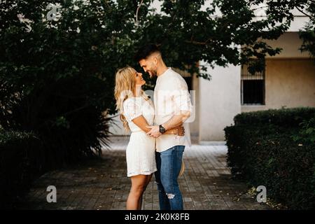 Side view of confident adult man and stylish woman in white dress looking at each other and smiling while standing and holding hands against green tre Stock Photo