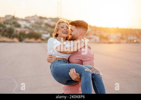 Confident man carrying on hands cheerful woman in trendy casual clothes while having fun on street against blurred town on hill during sunset Stock Photo