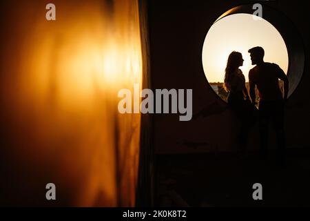 Anonymous couple silhouette in casual clothes looking at each other and laughing while sitting beside round window hole of modern building against blu Stock Photo