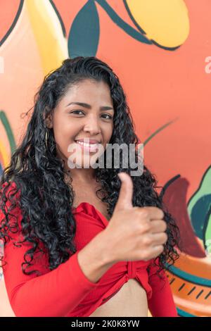 Cheerful young ethnic female with curly hair looking at camera while showing like gesture on colorful background Stock Photo