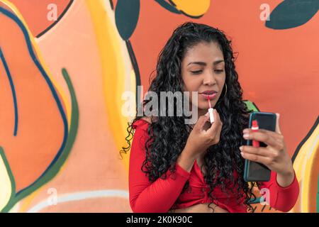 Young Latin American female with cellphone and curly long hair making up lips against graffiti wall Stock Photo